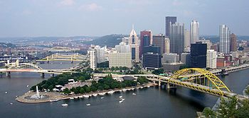 A view of a city nestled at the point where two rivers merge. There are yellow bridges crossing the rivers and a large fountain at the point where they meet. The city steps back from a park surrounding this fountain.