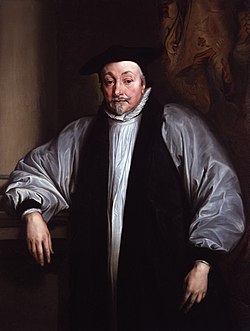 A man with a greying moustache and beard, wearing a white cassock and ruff with a black clerical scarf around his neck