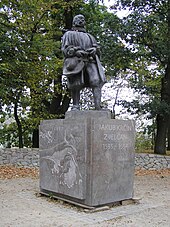 A statue of Jakub Krčín, depicted in dark metal, wearing voluminous clothes and a ruff around his neck. He is standing on a pedestal of grey stone, with a map of the left face of the Svět Pond, while on the front face has text chiselled into it, reading "Jakub Krčín Zjelčan 1535–1604". The picture is taken in summer, with the trees green and vibrant.