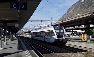White train on tracks with canopy-covered platforms on both sides and mountains to the rear