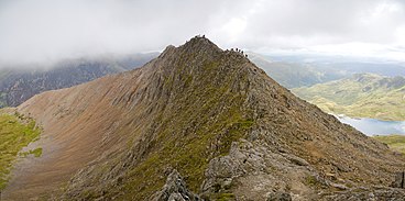 A sharp arête is seen obliquely; the sheer faces bear no vegetation and are made of noticeably reddish rock. Dozens of people can be seen clambering along the ridgeline.