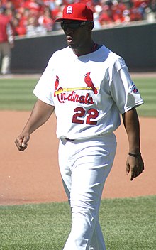 A dark-skinned man wearing a white baseball uniform with red trim, a red baseball cap, and sunglasses