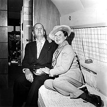 A black and white photograph of husband and wife Félix Bénitez Rexach and Lucienne D'Hotelle, seated on a bench inside their private airplane; Bénitez Rexach is wearing a dark pinstriped double-breasted suit finished with a spotted cravat and is reclining slightly; D'Hotelle is wearing a light wool pantsuit and matching hat, cocked at an angle, and is seated on the same bench with her legs up, reclining into her husband.