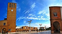 Two terrcota-coloured brick buildings, one of which has a clock, amongst a blue, clear sky