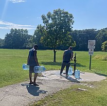 One man pumps water using a manual handle into a large empty water container, while another man waits with his own containers on a sunny day