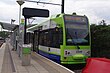 A tram platform with a shelter and ticket machine. A tram in green livery, with "West Croydon 2" written on the LED display is visible and a second set of tracks is visible in the top left corner.