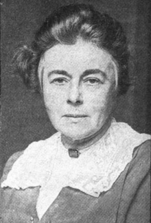 An older white woman, wearing a lace collar; her greying hair is dressed back from her face and off her shoulders