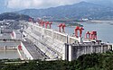 A photograph of a grey dam with red structures spaced across the top of it and with hilly terrain in the background all under a blue sky with white clouds