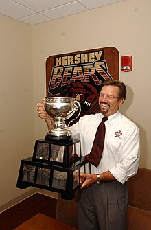 Doug Yingst, a brown-haired man wearing glasses and smiling, holds up the Calder Cup, a silver trophy atop two brown tiers. Behind Yingst is a poster with the Hershey Bears' logo.
