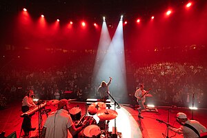 Four-man rock group performing on stage, spotlit, with row of darkened fans in the foreground