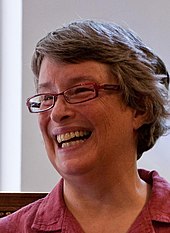 A bust-length photograph of a smiling woman with short grey hair wearing red glasses and a pink dress shirt while looking left and showing her teeth