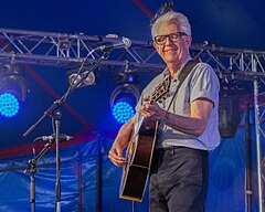 A white-haired man smiles as he strums his guitar on stage