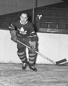 A man in full hockey gear skates toward the camera. He is in a dark jersey with a stylized maple leaf logo and is holding his stick left handed.