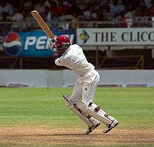 A man wearing white cricket clothes and maroon helmet plays a shot. He is standing on a cricket pitch, and the seating area of the ground is visible.