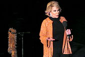 A woman with short blond hair wearing black except for a sparkly orange jacket with earrings. She is holding a microphone, and next to her is a microphone stand with an orange boa hanging from it.