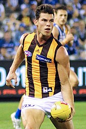 A male athlete with brown hair wearing a sleeveless guernsey and shorts prepares to handball a yellow football.
