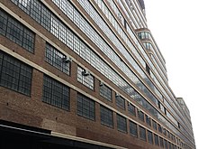 View of the facade from ground level. On the first story, mezzanine, and second story, the facade is divided vertically into multiple bays by brick piers. Above the second floor, the windows stretch across the facade in continuous horizontal ribbons.