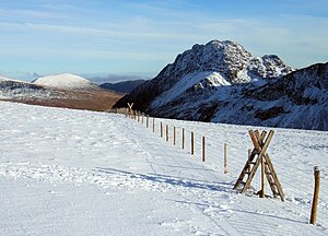 In the foreground, a gentle slope, covered with snow, is bisected by a wire fence with wooden stiles. Further back, a rugged mountain juts upwards.