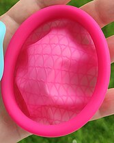 A menstrual disc/ring, made of silicone, bright pink. The rim has a round cross-section and is about 1cm thick; it forms an oval. The bowl is a crumpled, slightly translucent silicone membrane, with a latticework of slightly raised ridges in the shape of a tesselation of hearts.