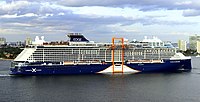 A blue-painted cruise ship is viewed from her starboard (right) side as she sails out of port; along the middle of the vessel is a tangerine sheltered platform that moves on a track on the starboard side