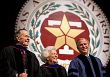 The head and shoulders of three people - an older man, an older woman, and a middle-aged man - wearing formal robes are shown in front of a large circular seal. On the outer edges of the seal the letters "XAS A...IVERSITY...87..." are visible; an inner band of leaves separates the letters from a block T superimposed with a star.