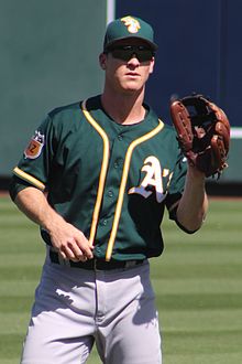 A man wearing a dark green cap and jersey with a white "A" on the chest and gray pants holds up his brown leather baseball glove preparing to catch a ball.