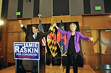 An older man and an older woman raise their hands together on a small stage in front of three flags