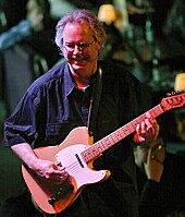 A man wearing glasses, playing a guitar and standing behind a cymbal.