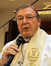 A bespectacled man wearing liturgical vestments and a pectoral cross holds a microphone in his right hand.