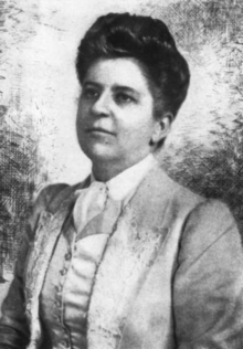 Lavinia Hartwell Egan, a white woman with dark hair in a bouffant updo, wearing a light-colored tailored suit with a collared white blouse