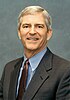 An older man is shown from the chest up, looking into the camera and smiling. He is wearing a gray suit, red patterned tie, and blue shirt. His hair is gray, and the background is a steely blue.