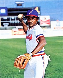 A man wearing a white baseball jersey with red and blue trim and a blue cap with a white "N" on the center stands with one arm back above his head preparing to throw a ball.