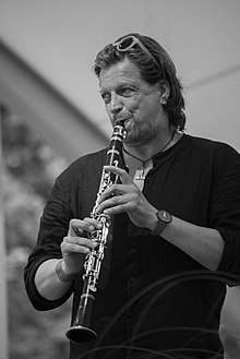 black and white photograph of a man with long hair playing a clarinet and looking to one side, probably on a stage