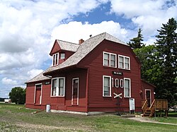 A small dirt path leads from the bottom right to the side of a red building and a five-step deck stairway at the end of which is a door. The side of the building is two stories, with two narrow windows on the bottom floor flanking the number 100, below which is a railroad crossing as a decoration and above which is a sign reading "Miami". Above these are two square windows. The building extends away to the left, with the steep roof pierced by a gable, beyond which is the roof of the single-storey freight shed. The walls consist of horizontal planks painted red, and the window frames are painted white. Behind the building are a few trees, with a cloudy sky in the background, and fronting the structure is green grass, patchy in some areas.