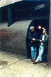 Two men wearing jackets and jeans, shaking hands, while standing in a passageway by the sidewalk of a brick building