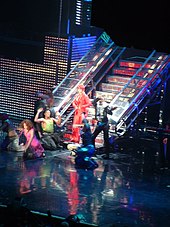 A woman wearing a red ensemble, standing in front of a metallic staircase around a group of dancers.