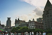 Pedestrians walk before a row of trees and a series of tall buildings. A blue sky overhead is obscured slightly by several clouds.