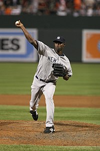 A man standing on the pitcher's mound wearing a gray baseball uniform with "New York" written across the chest in navy blue letters and a navy blue cap with a white "NY" on the front in the midst of pitching a baseball.