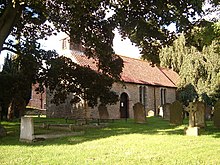 A stone church with a red tiled roof seen between trees in a churchyard. On the west gable is a bellcote.