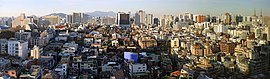 A daytime high-angle shot of a number of older mid-sized commercial and residential buildings on an incline. In the background, some larger office, department store, and church buildings are visible. In the very back, mountains and the North Seoul Tower are visible.