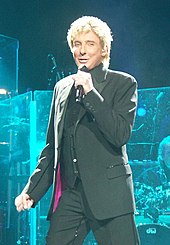 A blond-haired man wearing a dark suit, singing into a microphone