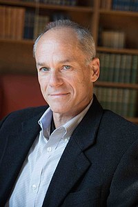 Marcelo Gleiser wearing a dark suit and light checkered shirt, gazing directly at camera with bookshelves in background