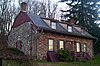 A small house made of reddish stone. It has a curved roof with three windows. Near the top of the roof the house's siding is maroon-colored wood.