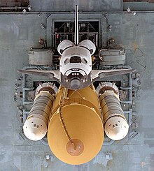 An overhead view of a spaceplane, coloured white on its topside and black on its underside, attached to a large orange tank, to which two slender white rockets are also attached. A gray platform supporting this stack serves as the background.