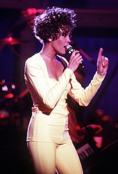A dark-skinned woman in a yellow top and pants singing into a microphone