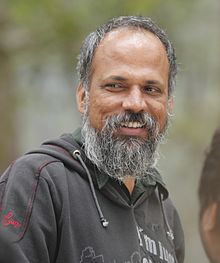 Amitabha Singh wearing a grey hoodie with lettering over an olive green shirt, grinning and looking right of camera