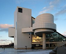 A grey tiled building rises over a lake, with a cylinder set on a narrow pole, and a sloping glass wall on one end.