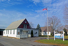 At left, a wooden one-story building with a tall gabled roof. The ground level is white with green trim; the upper level a dark reddish-brown, also with green trim. In the middle is a flagpole with the American flag; behind it is a white church partially obscured by a bare tree. At right is a blue and white sign with "Clermont" prominent on it