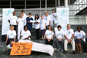 a group of people offering a petition. The group includes a person on a camp bed holding up a placard that says she is in bed 23 hours a day.