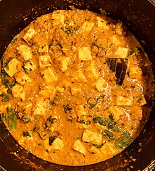 A gravy-based dish with many thick cheese cubes together with some vegetables and spices is shown. In the lower parts of the image, some coriander leaves have been added on top as a garnish.